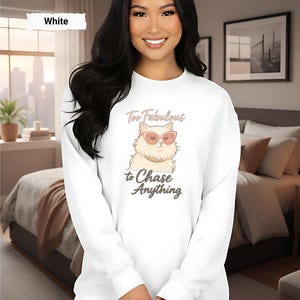 May include: White crew neck sweatshirt with a cartoon cat wearing sunglasses. The text on the shirt says "Too Fabulous to Chase Anything". The sweatshirt is a casual style.