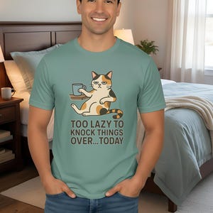 Könnte beinhalten: Hellgrünes T-Shirt mit einer Cartoon-Katze, die eine Kaffeetasse hält, und dem Text "TOO LAZY TO KNOCK THINGS OVER...TODAY". Das T-Shirt wird von einer Person in blauen Jeans getragen.