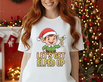 May include: White t-shirt featuring a cartoon elf holding a cocktail, wearing a green outfit and a red hat. The text "LET'S GET ELFED UP" is printed below the elf. The background includes a Christmas tree and festive decorations.