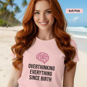 May include: A soft pink t-shirt with the text "OVERTHINKING EVERYTHING SINCE BIRTH" in black, along with a pink brain graphic. The shirt is worn by a person with long red hair, against a beach backdrop.