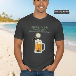 Puede incluir: Camiseta gris oscuro jaspeado con el texto "Looking For My Pot Of Gold (It's At the Bottom Of This Glass)". El gráfico muestra una lupa sobre una jarra de cerveza con un arcoíris y un pote de oro.