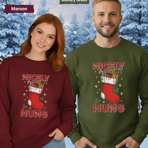 May include: Maroon and military green sweatshirts with a Christmas stocking graphic that reads "NICELY HUNG". The stocking is red with a white cuff and filled with gifts. The background is a snowy forest.