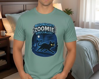 Pode incluir: Camiseta verde claro com um gráfico de um gato preto e as palavras "MIDNIGHT ZOOMIE SPECIALIST" em um design circular. O gato está correndo em um quarto escuro com uma janela. O modelo está usando jeans azul.