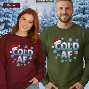 May include: Two sweatshirts, one maroon, one military green, with the text "COLD AF" in icy blue, snowflakes, and a Santa hat. A snowy forest scene forms the background. The sweatshirts are worn by two people.