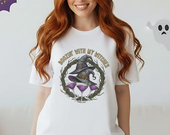 Puede incluir: Camiseta blanca con un diseño temático de Halloween. El diseño presenta un sombrero de bruja, copas de martini y el texto "Boozin' with my Witches" en una disposición circular. Decoraciones de murciélagos y fantasmas morados en el fondo.