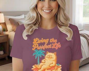 May include: Berry-coloured t-shirt featuring the text "Living the Purrfect Life" and a cartoon cat relaxing on a beach. The cat is orange and the palm tree is green. The model is wearing blue jeans.