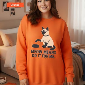 May include: Orange sweatshirt featuring a Siamese cat illustration with the text "MEOW MEANS DO IT FOR ME." The cat is depicted with a black face, paws, and tail, and a black collar. The sweatshirt is a solid orange color.