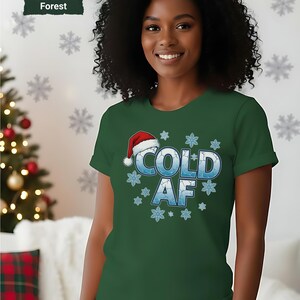 May include: Forest green t-shirt with the text "COLD AF" in blue, decorated with snowflakes and a Santa hat. The shirt is a festive winter holiday design.