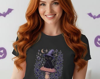 May include: Dark grey t-shirt with a cartoon black cat wearing a witch's hat, perched on a mushroom. The design includes a crescent moon and stars, surrounded by floral elements. The shirt is worn by a person with long red hair.