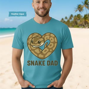 以下が含まれることがあります： ハート型のヘビのデザインと「SNAKE DAD」の文字が入った水色のTシャツ。柔らかい素材で作られています。「Heather Aqua」の文字が青い長方形に入っています。