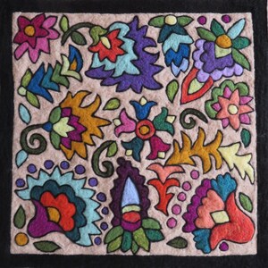 May include: A beige fabric square with a black border, featuring a colorful embroidered design of various flowers and leaves. The design includes shades of pink, purple, blue, green, yellow, and orange.