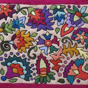May include: A colorful embroidered fabric square with a floral design. The fabric is beige with a pink border and features various flowers, leaves, and swirls in shades of red, orange, yellow, green, blue, purple, and pink.