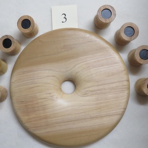 May include: A light-colored wooden game with a large circular base featuring a central hole, surrounded by eight cylindrical pieces with black centers. A small card with the number "3" is also present.