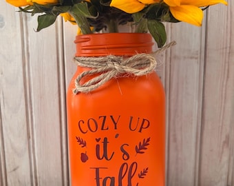 May include: A bright orange painted glass mason jar vase featuring a jute twine bow tied around the neck. The front of the jar displays the dark brown text "COZY UP it's Fall" with small leaf and acorn graphics. The jar is filled with yellow sunflowers.