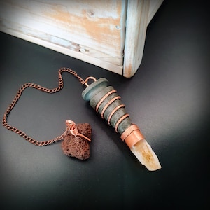 May include: A pendulum featuring a tapered dark grey stone wrapped in copper wire, attached to a copper chain. A small, porous reddish-brown volcanic rock is also attached to the chain, and a translucent yellow crystal point is set at the base.