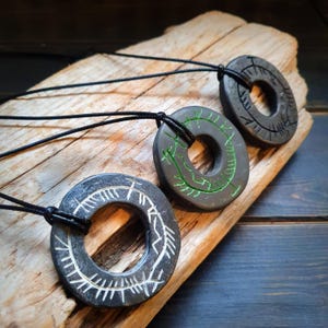 Peut inclure: Trois pendentifs en pierre gris foncé avec des motifs circulaires et des cordons noirs. Un pendentif a des marques blanches, un autre des marques vertes et le troisième des marques gris foncé. Les pendentifs sont exposés sur un morceau de bois flotté.