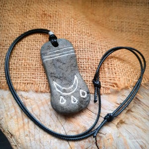 Könnte beinhalten: Grauer Steinanhänger mit einer schwarzen Kordelkette. Der Anhänger hat eingravierte weiße Linien und ein Halbmond-Design mit drei Diamantformen. Die Halskette ist geknotet und liegt auf einer Holzoberfläche.