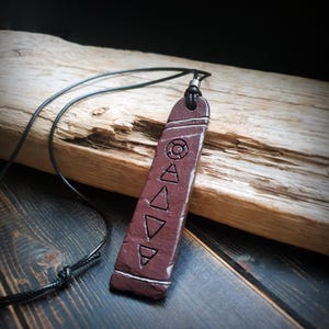 Puede incluir: Un collar con colgante granate con un cordón negro. El colgante presenta formas geométricas y líneas grabadas en blanco. El collar se exhibe sobre madera flotante, con un fondo de madera oscura.