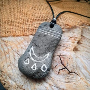 Può includere: Ciondolo in pietra grigia con cordino nero. Il ciondolo presenta un disegno intagliato con una luna crescente e tre forme a goccia. Due linee orizzontali sono intagliate vicino alla parte superiore della pietra.