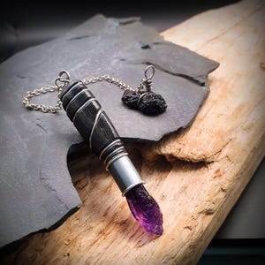 May include: A pendant featuring a dark, cylindrical, wire-wrapped casing attached to a silver-toned chain. A translucent, deep purple, pointed crystal extends from the bottom of the casing. Two small, dark, porous stones are attached to the chain.