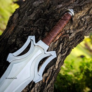 May include: A silver and brown sword with a leather-wrapped handle rests against a tree trunk. The blade is silver with a decorative design. The handle is brown leather. The sword is a fantasy-themed item.