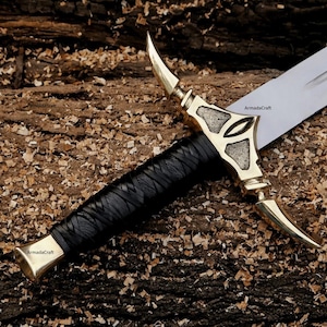 May include: A close-up of a decorative sword with a black leather-wrapped handle and a gold-colored hilt. The hilt features a stylized design with curved elements. The blade is silver and partially visible. The sword rests on a bed of wood shavings.