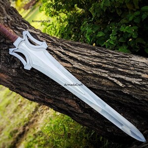 May include: A silver fantasy sword with a brown leather-wrapped handle rests on a tree trunk. The blade is long and has a distinctive shape with decorative cutouts near the hilt. The sword is a prop or collectible item.