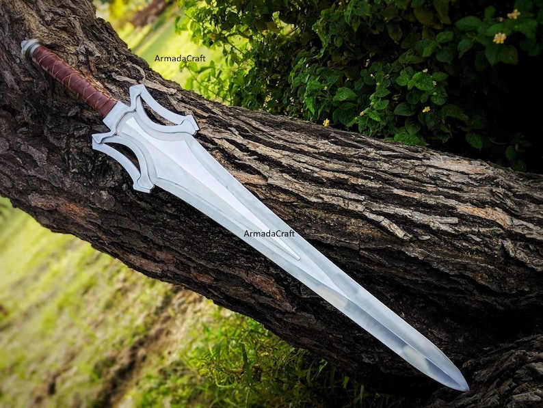 May include: A silver fantasy sword with a brown leather-wrapped handle rests on a tree trunk. The blade is long and has a distinctive shape with decorative cutouts near the hilt. The sword is a prop or collectible item.