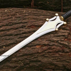 May include: A silver-colored sword with a decorative hilt and a black handle. The hilt has gold-colored accents and a cream-colored center. The sword is lying on a brown, textured surface.