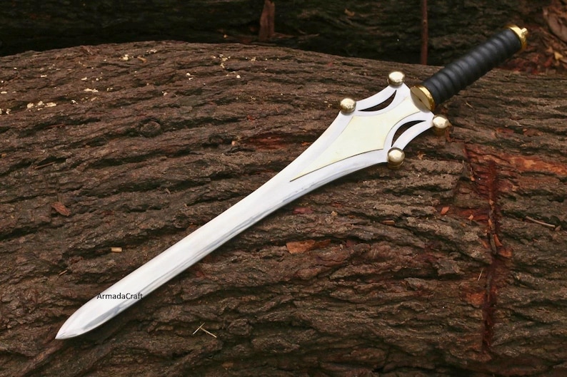 May include: A silver-colored sword with a decorative hilt and a black handle. The hilt has gold-colored accents and a cream-colored center. The sword is lying on a brown, textured surface.