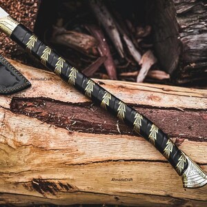 May include: A handcrafted axe with a black handle adorned with gold leaf patterns. The axe head is silver, and a black leather sheath is visible. The axe rests on a weathered wooden surface, showcasing the craftsmanship.
