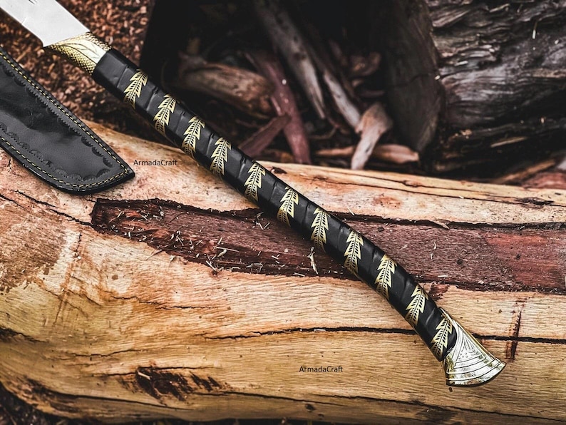 May include: A handcrafted axe with a black handle adorned with gold leaf patterns. The axe head is silver, and a black leather sheath is visible. The axe rests on a weathered wooden surface, showcasing the craftsmanship.