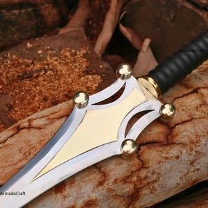 May include: A close-up of a decorative sword with a silver blade and a gold-colored hilt. The handle is wrapped in black leather. The hilt features gold accents and four gold spheres. The text "ArmadaCraft" is visible.