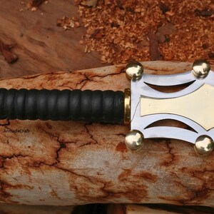 May include: Close-up of a decorative dagger with a black leather grip and a silver and gold blade. The handle has gold accents. The dagger is resting on a piece of wood.