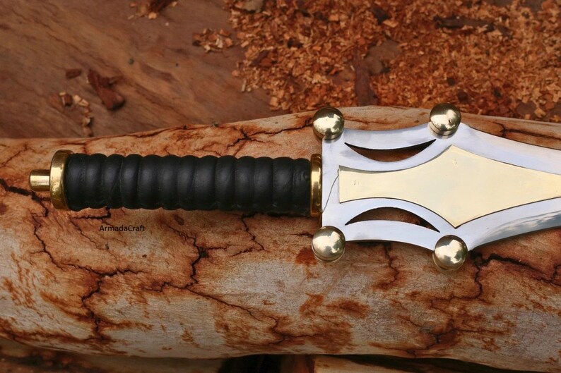 May include: Close-up of a decorative dagger with a black leather grip and a silver and gold blade. The handle has gold accents. The dagger is resting on a piece of wood.