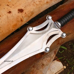 May include: A silver and black sword with a double-edged blade. The hilt is wrapped in black leather, and the guard features decorative cutouts and silver ball accents. The sword rests on a wooden surface.