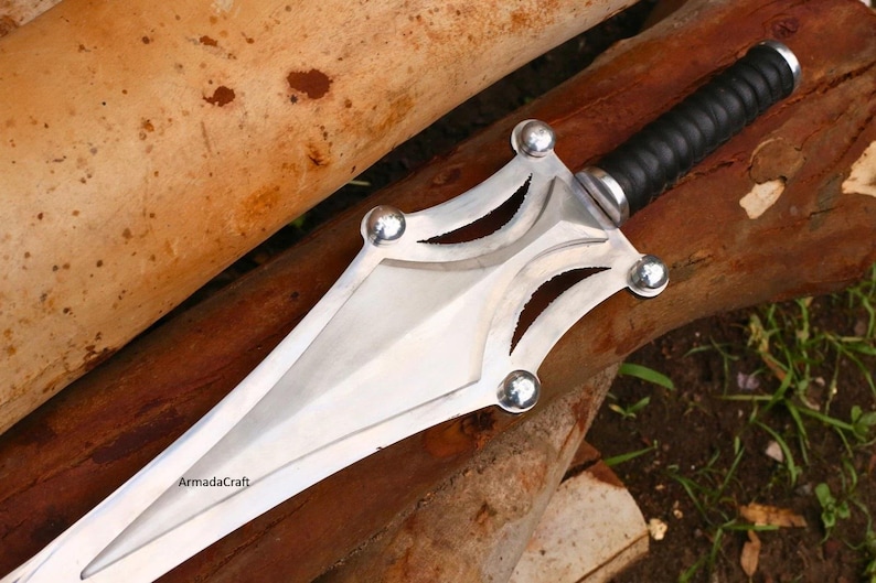 May include: A silver and black sword with a double-edged blade. The hilt is wrapped in black leather, and the guard features decorative cutouts and silver ball accents. The sword rests on a wooden surface.