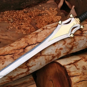 May include: A decorative sword with a silver blade and a gold and black handle. The sword features a gold-colored guard with decorative cutouts and gold accents. The handle is wrapped in black material. The sword is resting on a wooden surface.