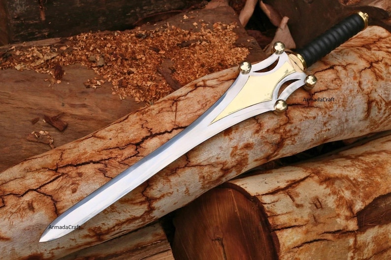 May include: A decorative sword with a silver blade and a gold and black handle. The sword features a gold-colored guard with decorative cutouts and gold accents. The handle is wrapped in black material. The sword is resting on a wooden surface.