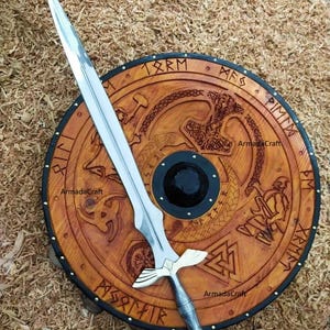 May include: A wooden shield with intricate carvings and a long, silver sword. The shield is round, with a dark border and a central black circle. The sword has a detailed hilt and a sharp blade. The shield and sword are displayed on a bed of wood shavings.