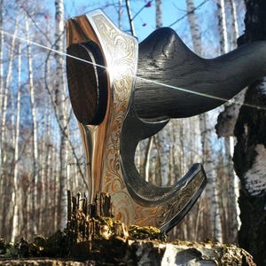 May include: A close-up of a decorative axe with a silver and gold-toned head and a dark wooden handle. The axe head features intricate engraved patterns. The axe is resting on a mossy, wooden surface, with a blurred forest background.