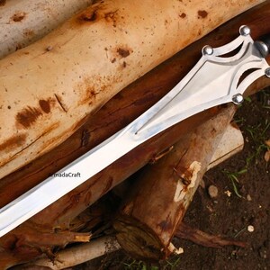 May include: A silver sword with a black handle rests on a bed of brown logs. The sword has a wide, polished blade and a decorative hilt with silver accents. The handle is wrapped in black material.