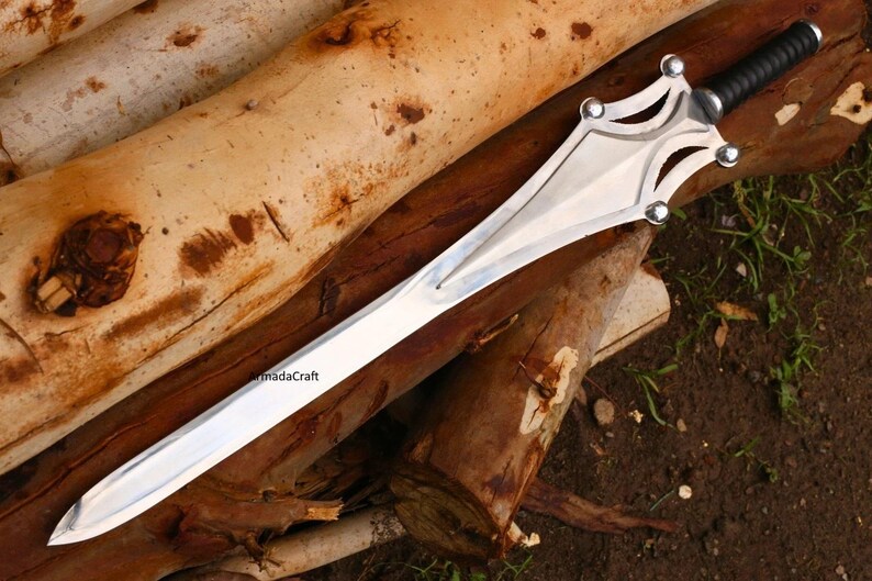 May include: A silver sword with a black handle rests on a bed of brown logs. The sword has a wide, polished blade and a decorative hilt with silver accents. The handle is wrapped in black material.