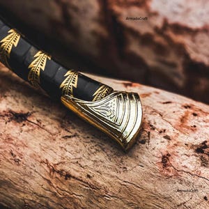 May include: Close-up of a decorative dagger with a black handle adorned with gold leaf patterns. The gold-colored metal guard features intricate engraved designs. The dagger rests on a textured wooden surface.