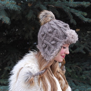 Puede incluir: Una mujer lleva un gorro de punto gris con un pompón de piel sintética y orejeras. El gorro tiene un ribete de piel sintética blanca alrededor de las orejeras. También lleva un chaleco de piel sintética blanca.