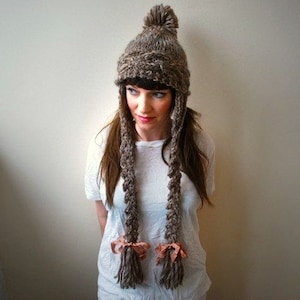 May include: A woman wearing a brown knit hat with long ear flaps and a pom-pom on top. The hat has two long, brown tassels tied with pink ribbons at the ends.