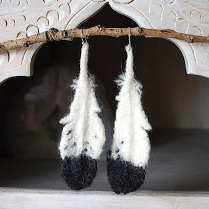 Puede incluir: Un par de pendientes de plumas blancas y negras colgando de una rama. Las plumas son blancas con puntas negras y están hechas de fieltro.