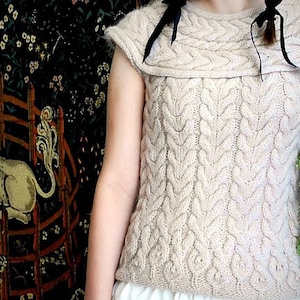 Puede incluir: Un top de punto sin mangas de color crema con un patrón de punto de cable y un escote ancho y doblado. El top lo lleva una persona con una cinta negra atada al cuello.