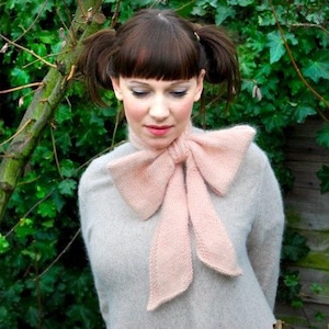 May include: A woman with brown hair and bangs wearing a light grey sweater with a large pink bow tied around the neck. The woman is standing in front of a green leafy background.