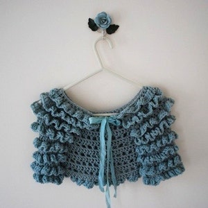 Pode incluir: Um bolero de crochê azul claro com bordas franzidas, pendurado em um cabide branco. Uma fita azul combinando está amarrada na frente. Uma flor azul decora a parede acima do cabide.